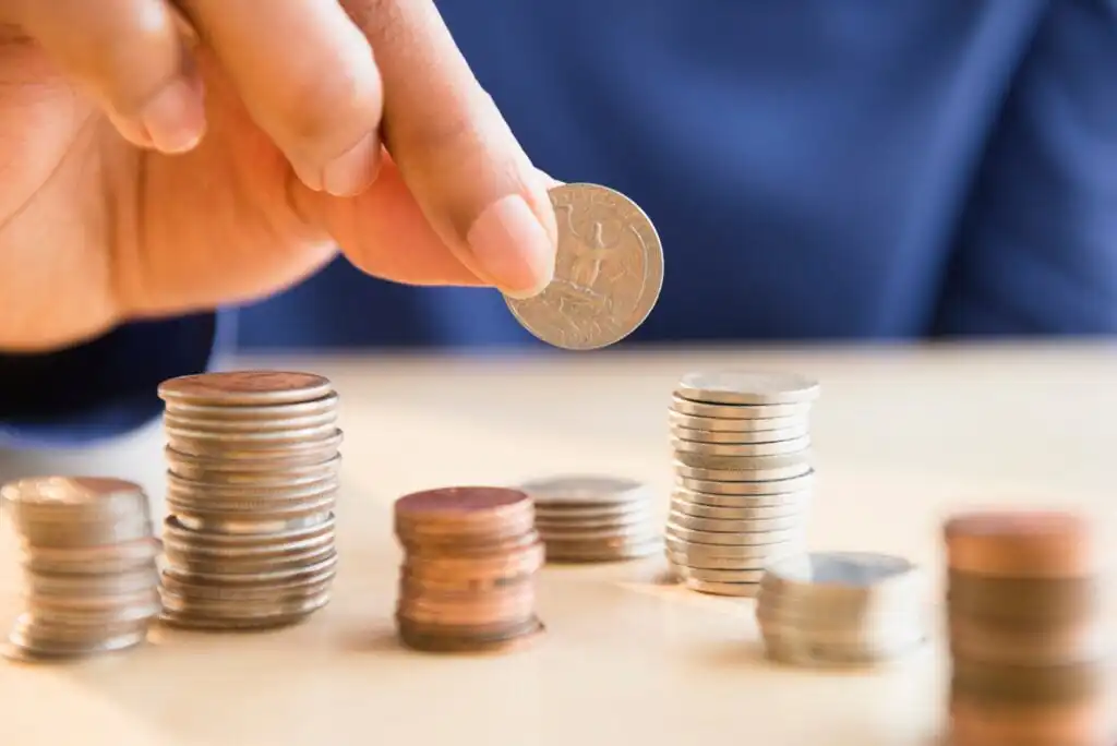 A hand holds a coin above several small stacks of coins arranged on a light-colored surface, suggesting counting or saving money. The background is slightly blurred.