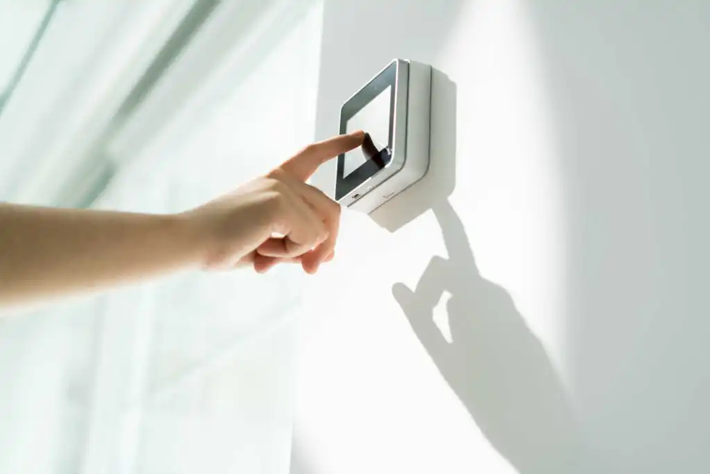 A hand is touching the screen of a wall-mounted smart thermostat, casting a shadow on the white wall beside it.