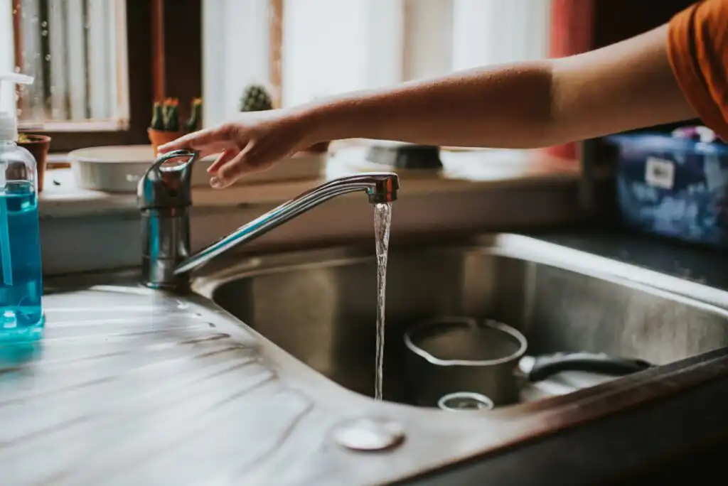 A person’s arm reaches to turn off the faucet above a stainless steel kitchen sink with water running, dishes and soap visible nearby.