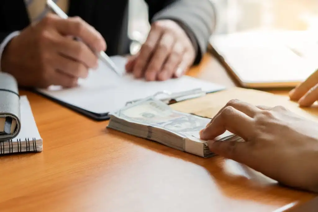 Two people sit at a desk; one is writing on a clipboard while the others hand reaches for a stack of US hundred-dollar bills. A brown envelope and documents are also on the table.