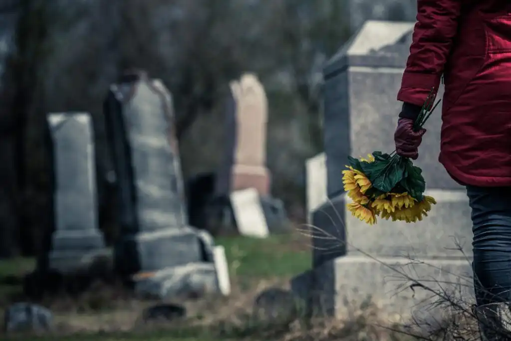 A person in a red coat holds a bouquet of yellow flowers while standing in a graveyard with several old, weathered headstones in the background.