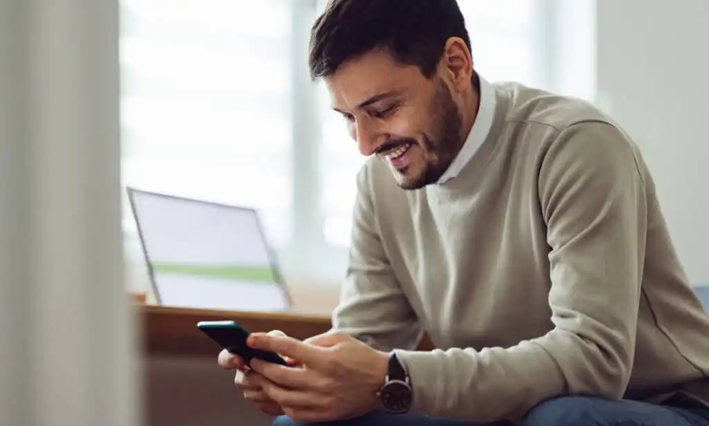 A man with short dark hair and a beard smiles while looking at his smartphone. He is wearing a light sweater and jeans, sitting indoors near a blurred laptop on a desk in the background.