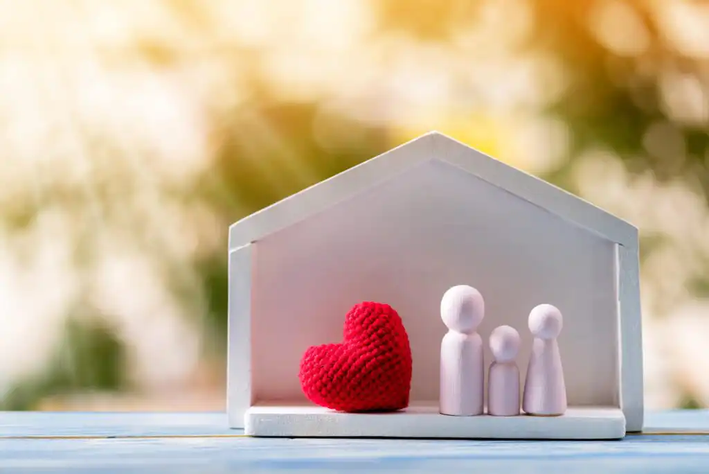 A white wooden house frame with three wooden figures representing a family and a red knitted heart inside, set on a blue surface with a blurred outdoor background.