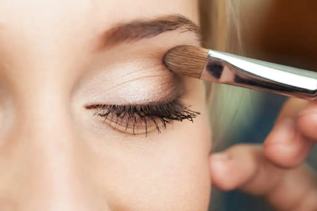 A close-up of a persons closed eye as they apply shimmery beige eyeshadow to their eyelid with a makeup brush. Their eyelashes are coated with mascara, and their eyebrow is neatly groomed.