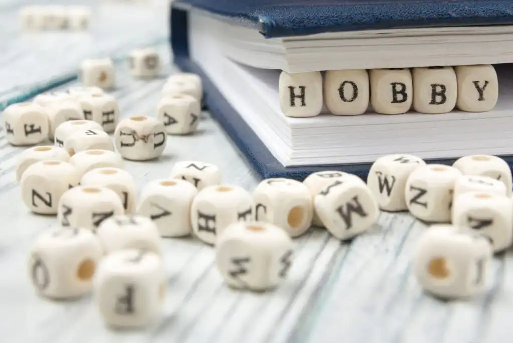 White beads with black letters are scattered on a light wooden surface, with some beads spelling HOBBY tucked between the pages of a closed blue book.