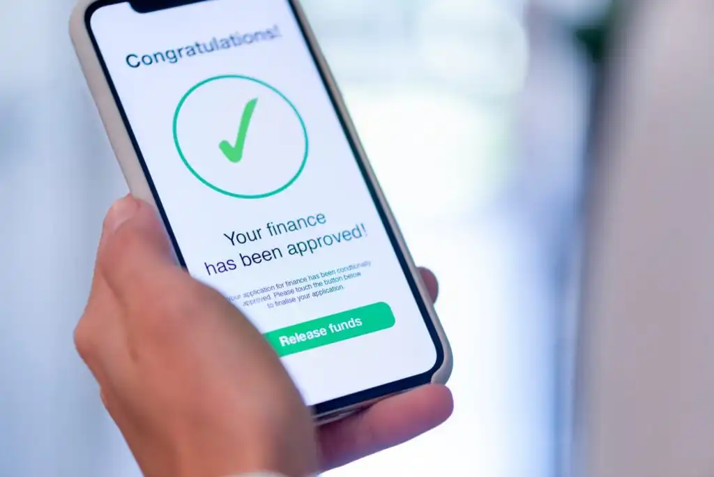A hand holds a smartphone displaying a screen that reads, Congratulations! Your finance has been approved! with a green check mark and a Release funds button.