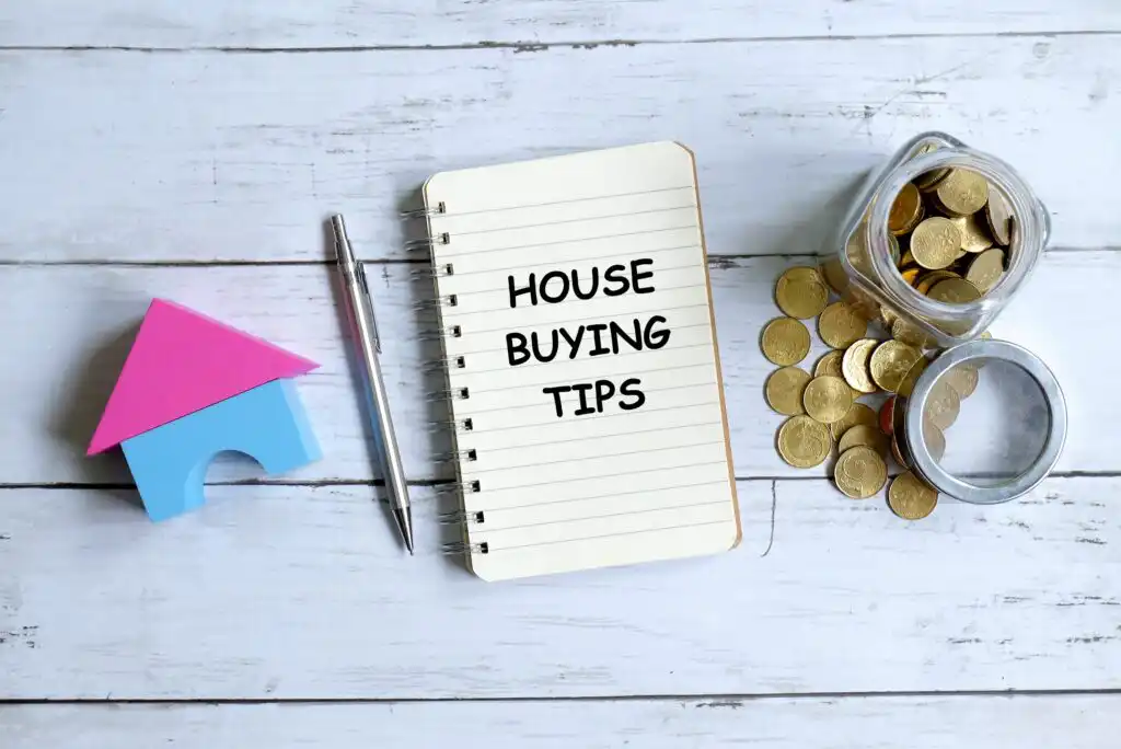 A notebook with HOUSE BUYING TIPS written on it, a pen, a small toy house, and a jar of gold coins are arranged on a white wooden surface.