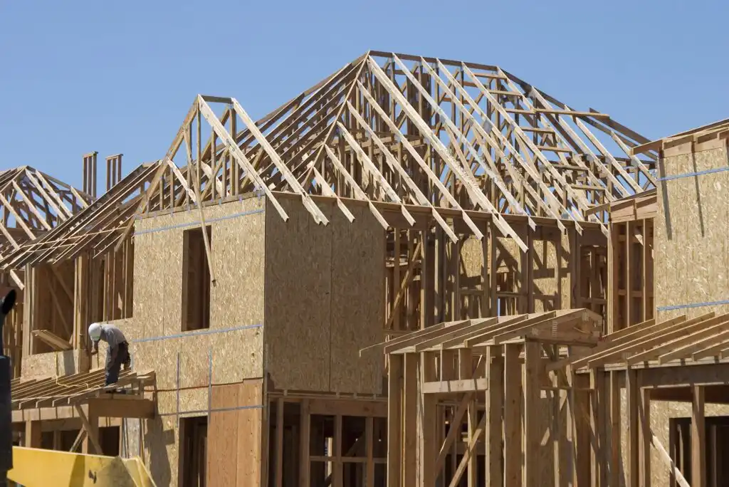 A house under construction with wooden framing and exposed plywood walls. A worker is on the left side, working on the structure. The sky is clear and blue.