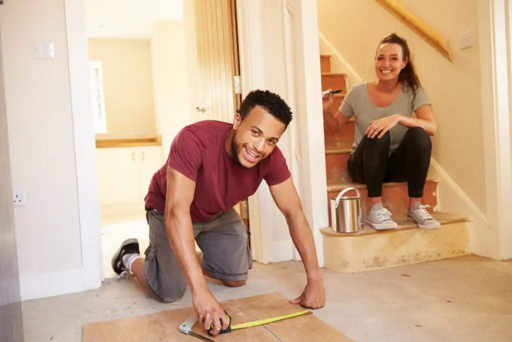 A man measures wood with a tape measure on the floor of a home under renovation, smiling at the camera. A woman sits on the stairs behind him, also smiling, holding a paintbrush.