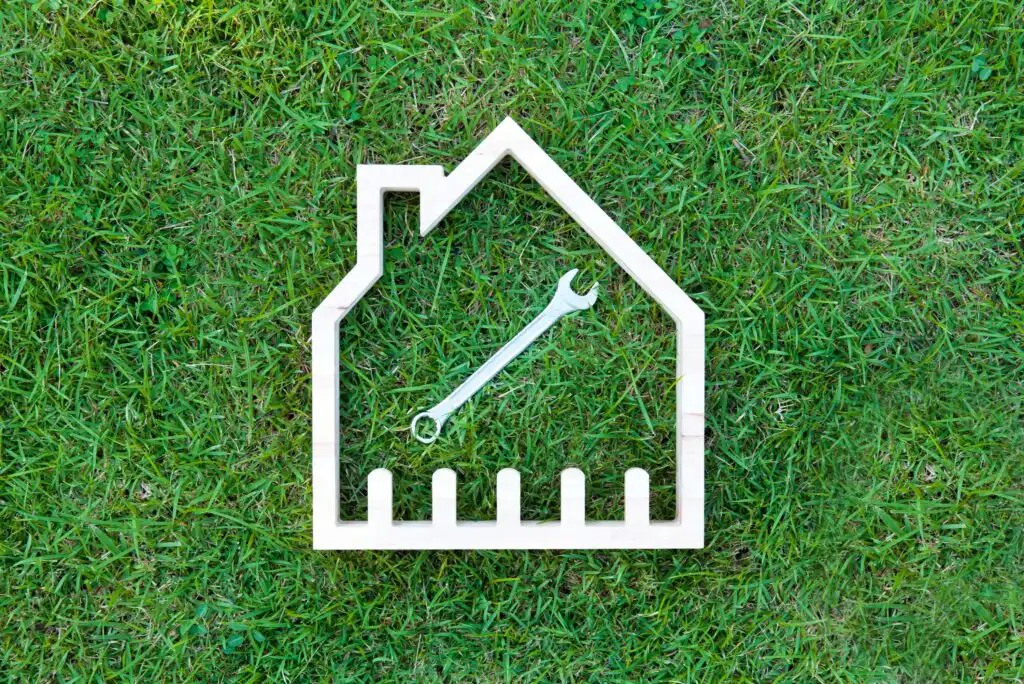 A wooden house-shaped outline with a metal wrench inside is placed on green grass, symbolizing home repairs or maintenance.