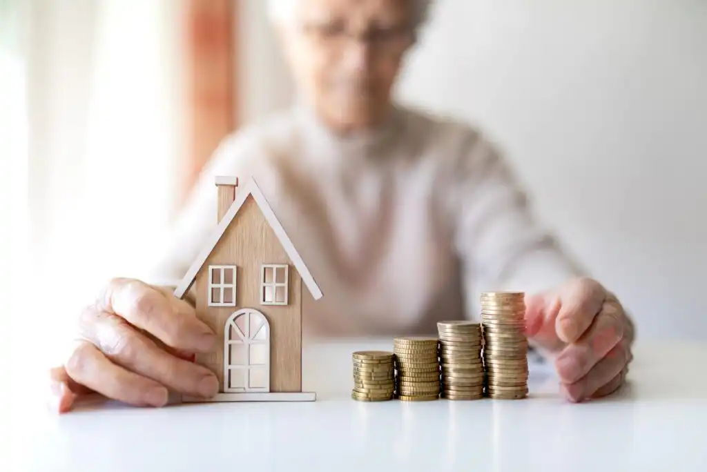 An elderly person holds a small wooden house model and stacks of coins arranged in ascending order on a white table, symbolizing savings, investment, or retirement planning.
