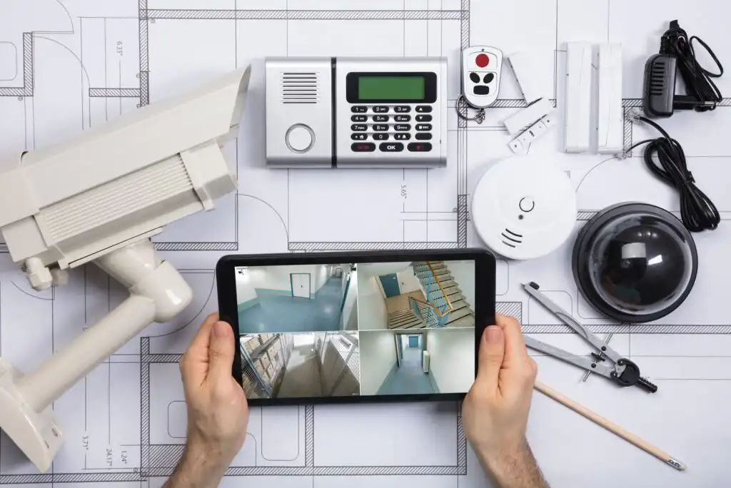 A person holds a tablet showing security camera feeds over blueprints, surrounded by security equipment such as a CCTV camera, alarm keypad, motion sensors, smoke detector, dome camera, and electrical components.