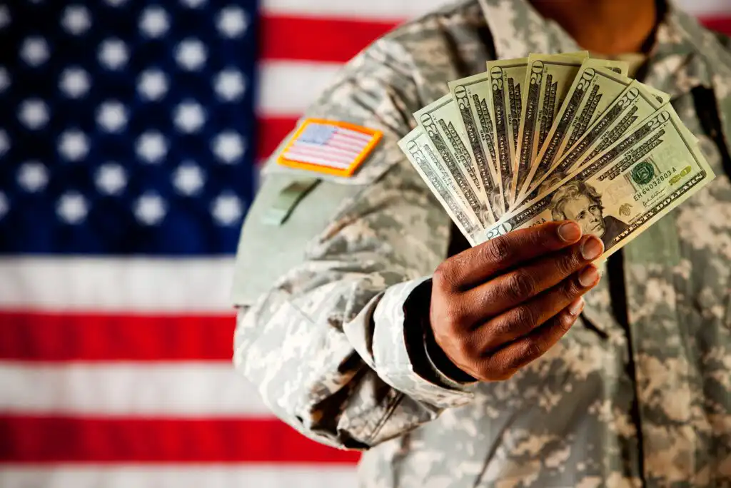 A person in military uniform holds out a fan of U.S. dollar bills, with an American flag in the blurred background.