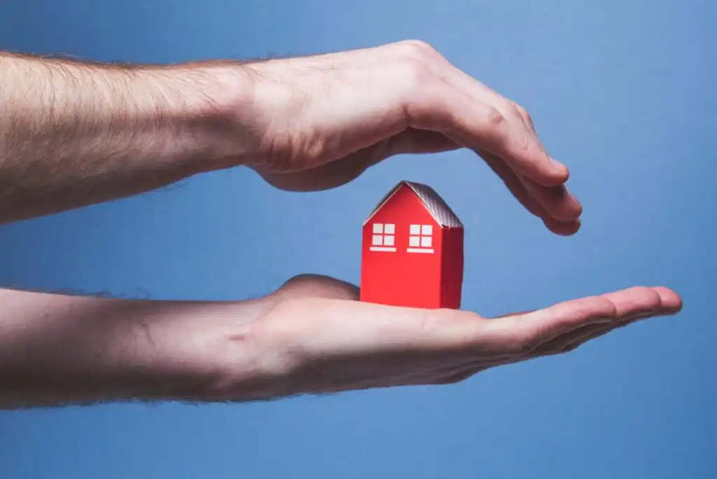 A pair of hands gently cradles and shelters a small red house model against a light blue background, symbolizing home protection or insurance.