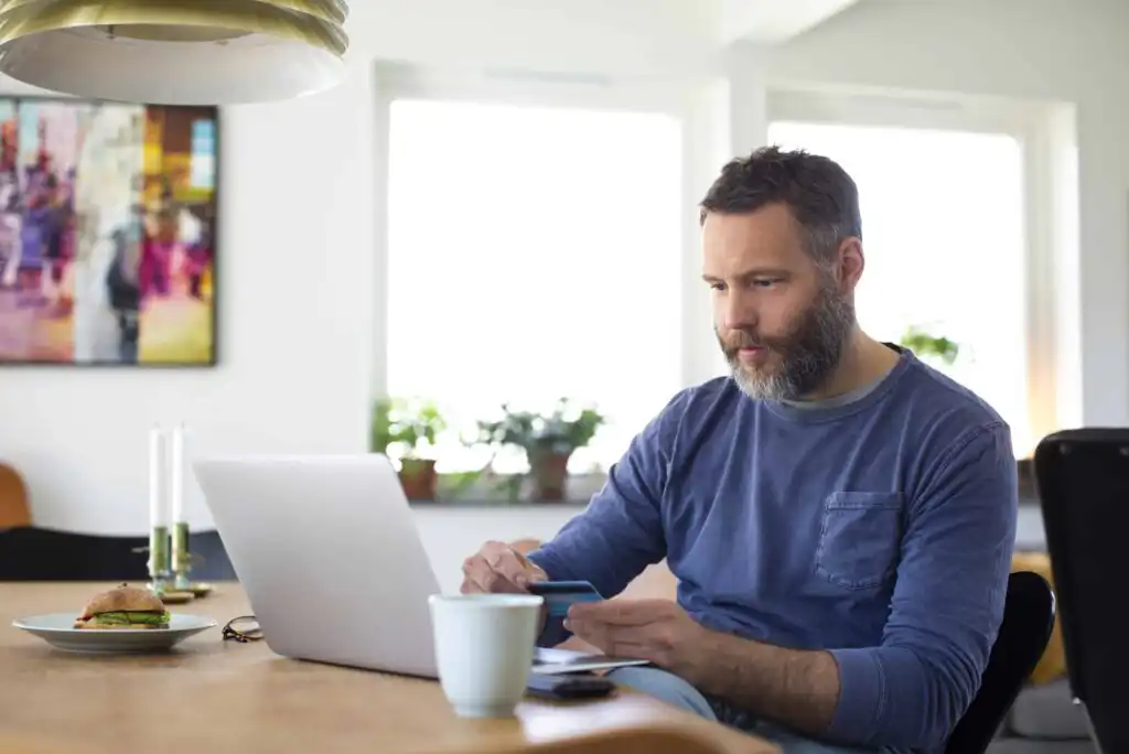 A man with a beard sits at a wooden table using a laptop and holding a credit card. There is a coffee mug and a sandwich in front of him. The room is bright with large windows and several plants in the background.