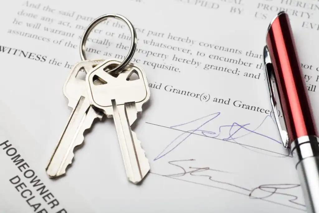 Two silver house keys on a keyring rest on a signed legal document, next to a red and silver pen. The document is related to a homeowner declaration or property agreement.