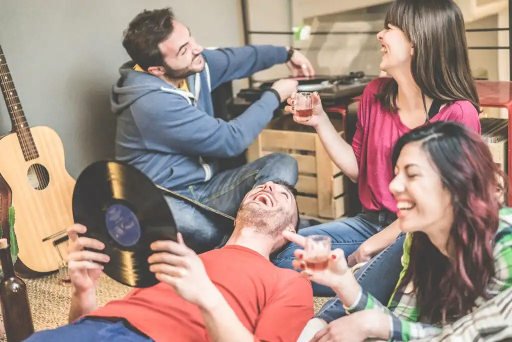 Four friends sit on the floor, laughing and enjoying drinks together. One person holds a vinyl record while another uses a turntable. A guitar rests against the wall in the background, giving a cozy, musical atmosphere.