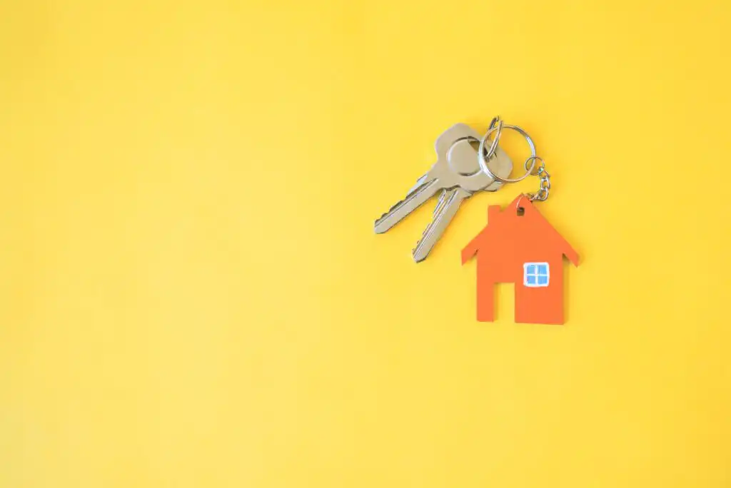 Two silver keys attached to a keyring with an orange house-shaped keychain with a blue window, all placed on a bright yellow background.