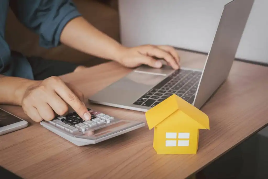 A person uses a laptop and presses buttons on a calculator at a desk, with a small yellow paper house model nearby, suggesting financial planning or calculating home expenses.