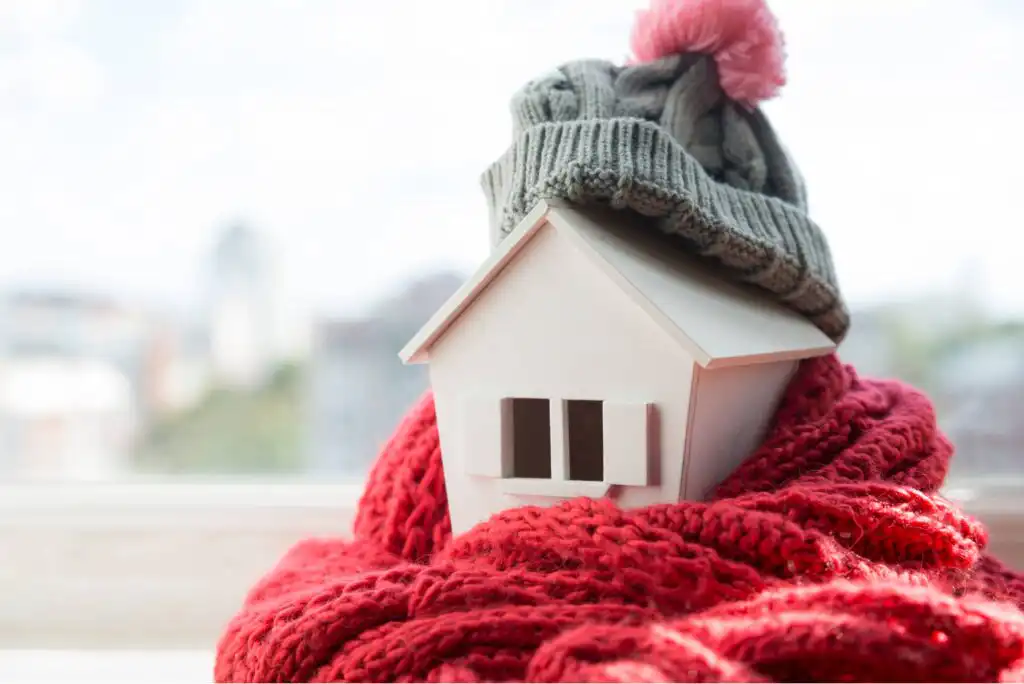 A small model house wrapped in a red knitted scarf and topped with a gray winter hat with a pink pom-pom, symbolizing warmth and home insulation.