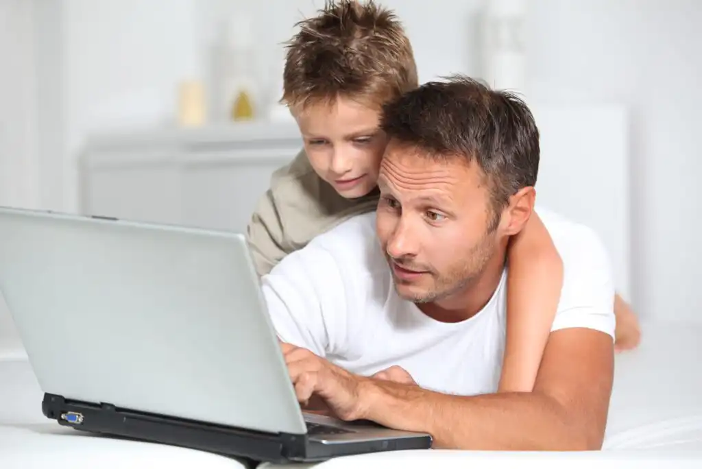 A man lying on a bed uses a laptop while a young boy leans over his shoulder, watching the screen. Both appear focused and engaged with the computer in a bright, home setting.