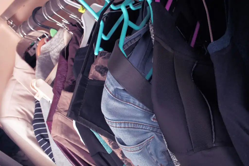 A close-up view of clothes hanging in a closet, including denim jeans, dark tops, and various garments on plastic hangers of different colors.