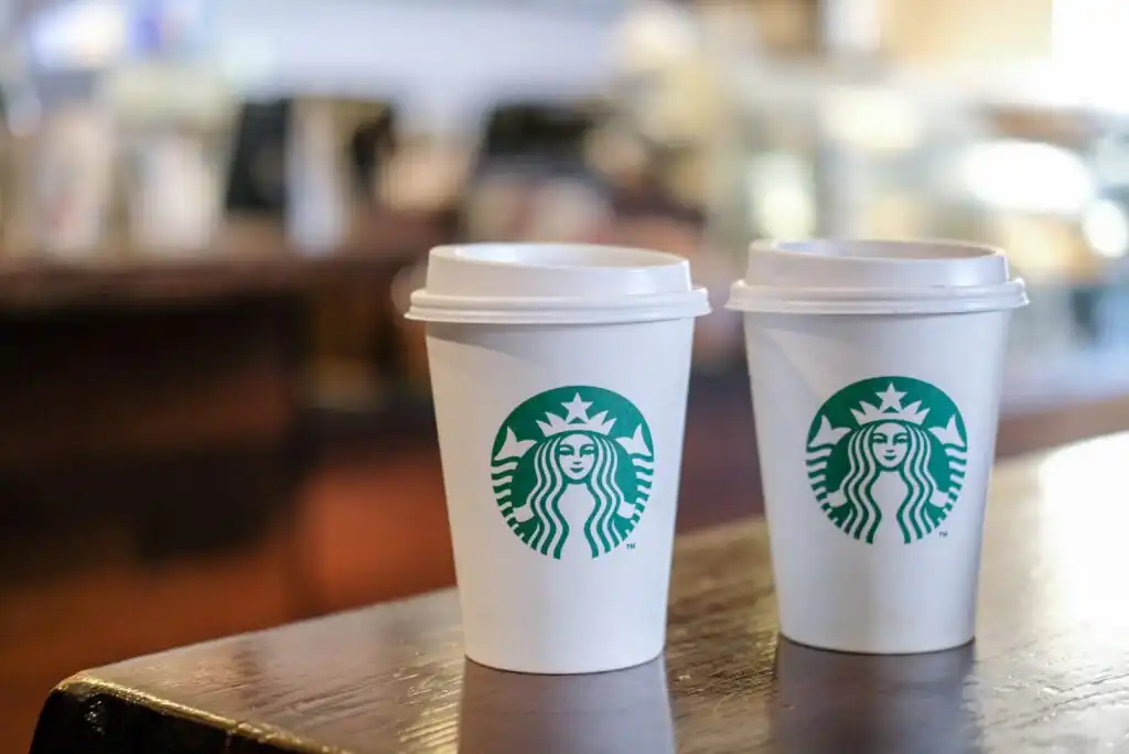 Two Starbucks coffee cups with lids sit side by side on a wooden table, with a blurred café background.