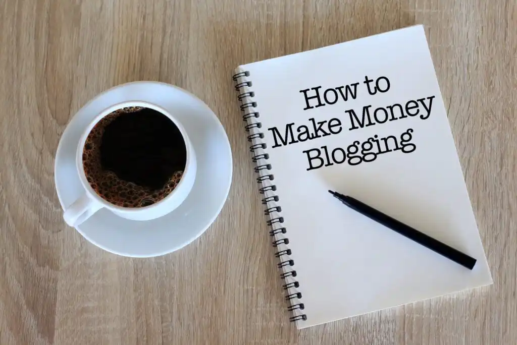 A cup of coffee in a white saucer sits next to an open spiral notebook with the words How to Make Money Blogging written on the page. A black pen rests on the notebook, all on a wooden table.