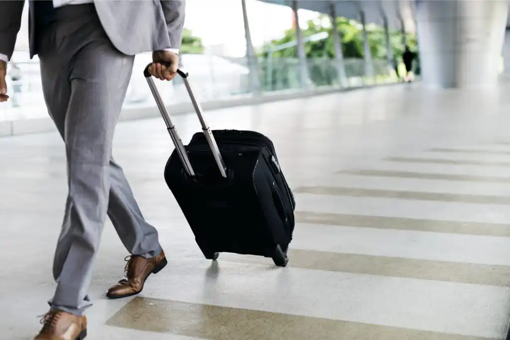 A person in a gray suit and brown shoes is walking indoors, pulling a black rolling suitcase along a striped floor in a modern, glass-walled building.