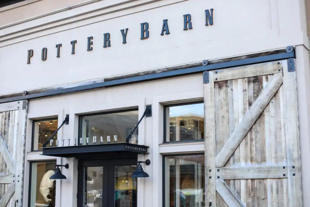 The exterior of a Pottery Barn store, featuring large windows, rustic wooden sliding doors, black light fixtures, and bold “Pottery Barn” signage above the entrance.