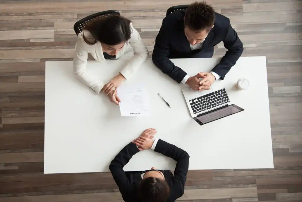 Top-down view of two people in business attire sitting across a desk from a candidate during a job interview. A resume, pen, and laptop are on the white table. All have hands folded or holding papers.
