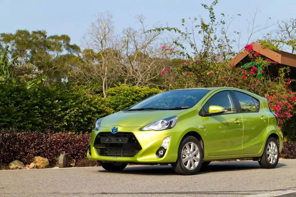 A bright green Toyota Prius C hatchback is parked on a paved road, surrounded by greenery, flowering bushes, and trees under a clear blue sky.