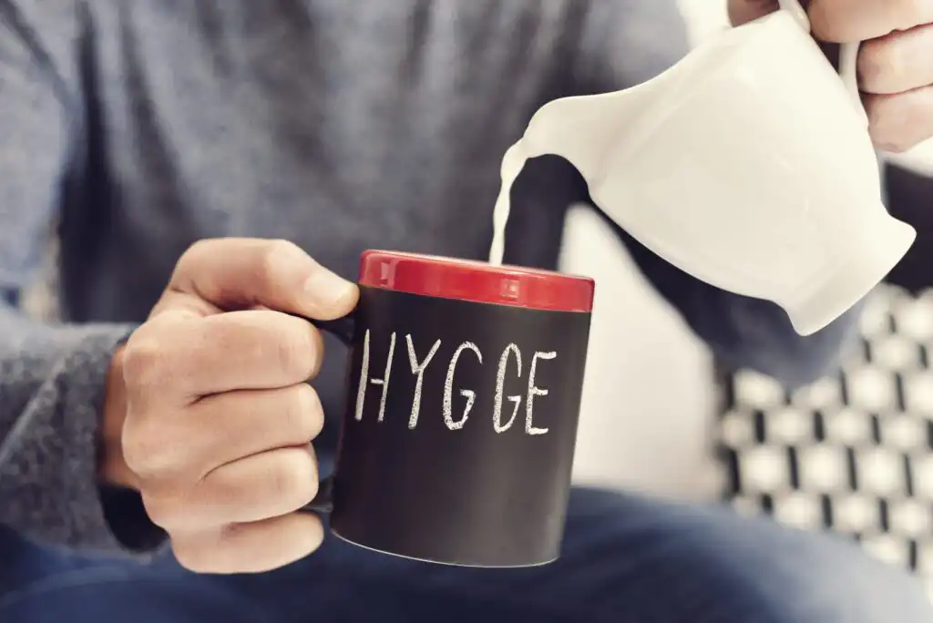 A person pours milk from a white pitcher into a black mug with a red rim and the word HYGGE written on it, creating a cozy atmosphere.