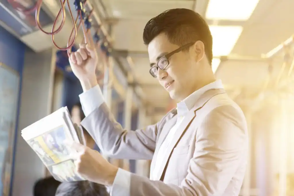 A man wearing glasses and a light-colored suit stands on a train, holding onto an overhead strap with one hand and reading a newspaper with the other, smiling in the bright, sunlit interior.