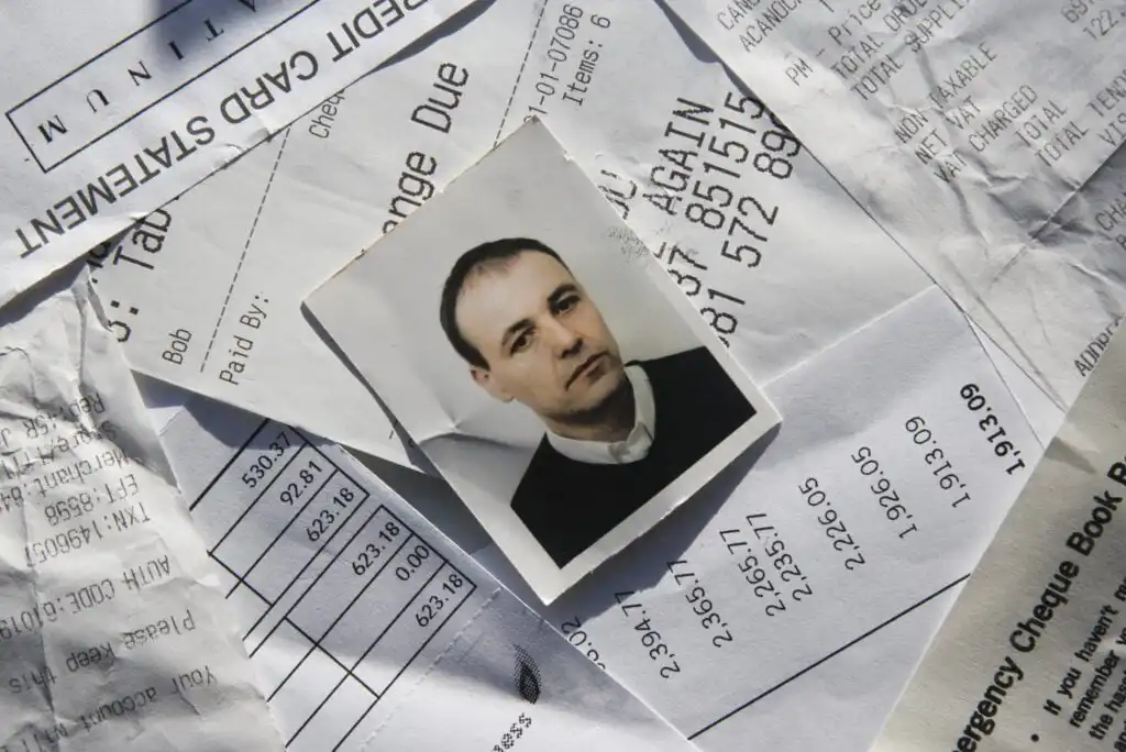 A small black-and-white portrait photo of a man lies on top of various receipts and financial documents scattered across a flat surface.