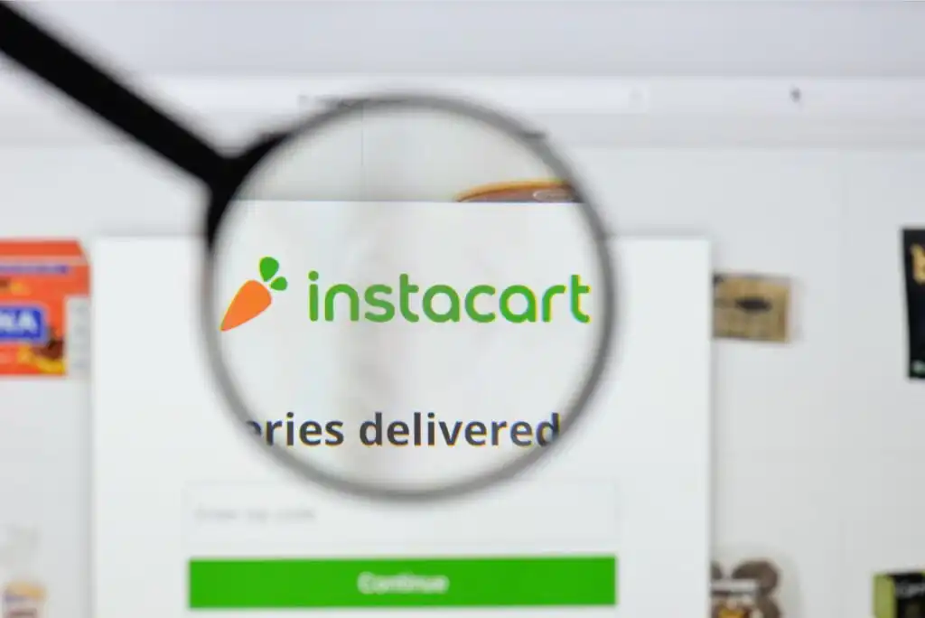 A magnifying glass focuses on the Instacart logo and part of its website, showing the green and orange logo with the word instacart and blurred grocery items in the background.