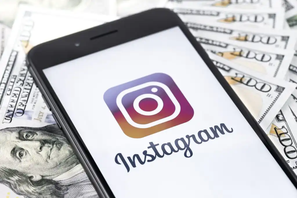 A smartphone displaying the Instagram logo and name rests on top of scattered US hundred-dollar bills.