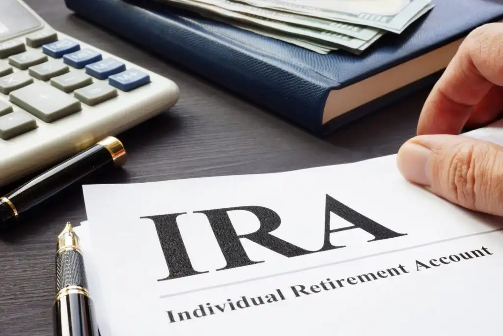 A paper labeled IRA Individual Retirement Account on a desk with a pen, calculator, a closed notebook, and a stack of cash visible in the background. A hand is holding the paper.