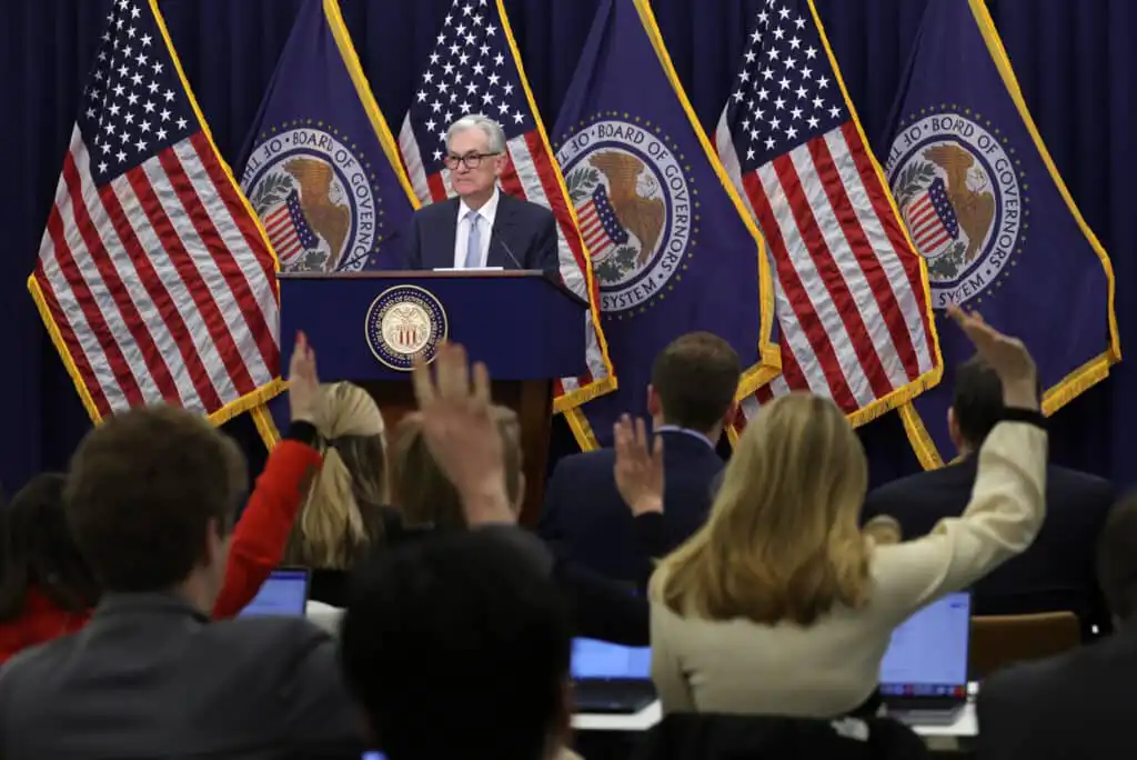 A man stands at a podium labeled Federal Reserve with several U.S. flags and official emblems behind him. He faces an audience with raised hands, some using laptops, appearing to ask questions.
