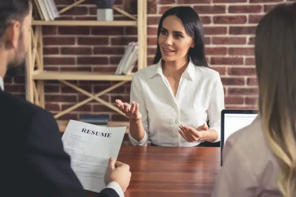 A woman in a white shirt sits at a table, speaking and gesturing during a job interview. Two people face her, one holding a resume. A laptop and shelves with books are visible in the background.