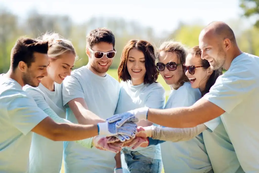 A group of seven smiling people wearing matching light blue shirts stand outdoors, stacking their hands together in a show of teamwork and unity.