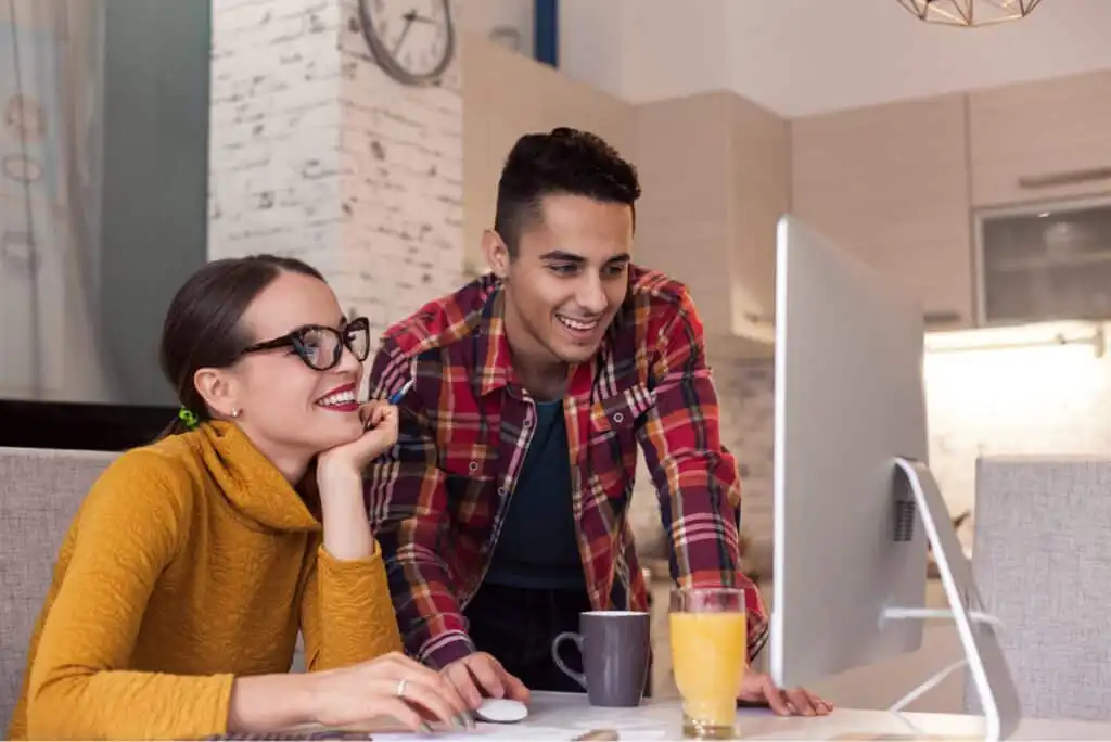 A woman in glasses and a yellow sweater and a man in a red plaid shirt smile while looking at a computer screen in a modern kitchen; a mug and a glass of orange juice are on the table.