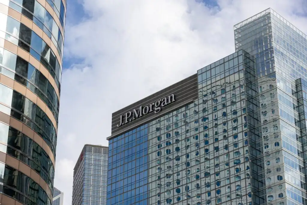 Glass skyscraper with the J.P. Morgan logo near the top, set against a cloudy sky, with reflections on the buildings glass windows and other tall buildings partially visible nearby.