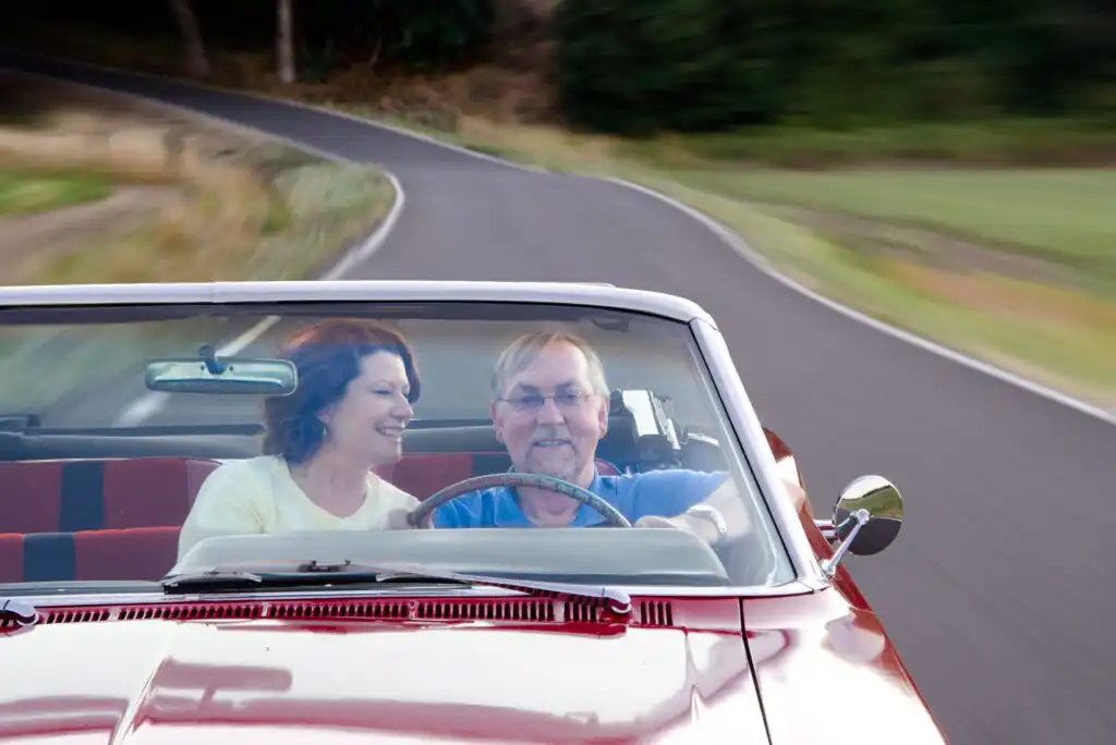 An older couple smiles and enjoys a drive in a red convertible car on a winding road, surrounded by greenery and blurred motion, suggesting movement and a carefree atmosphere.