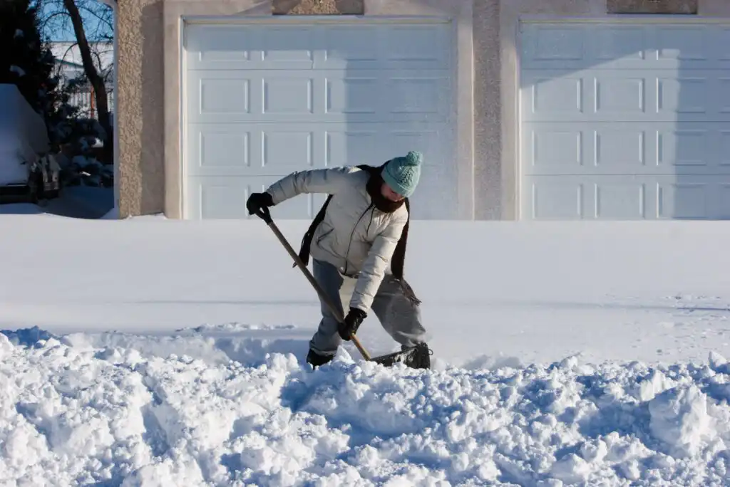 A person wearing winter clothes shovels snow from a driveway in front of a house with double garage doors on a sunny day.