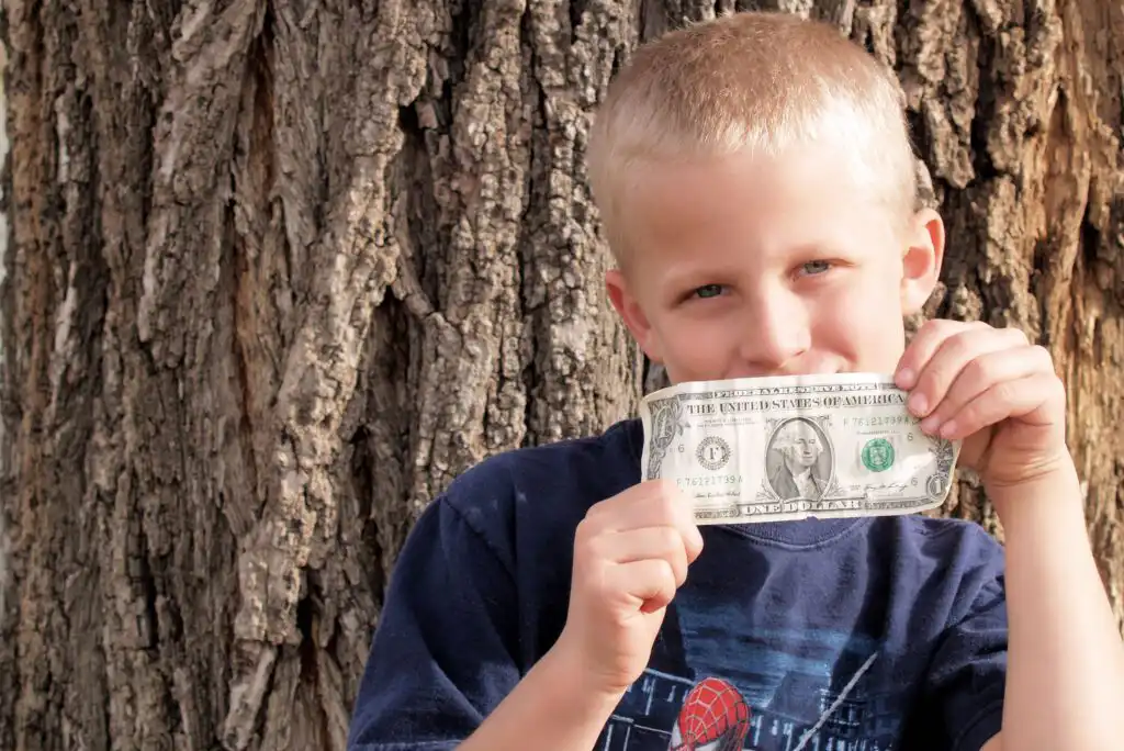 A young boy with short blond hair holds up a one-dollar bill and smiles while standing in front of a large tree trunk, wearing a dark blue t-shirt.