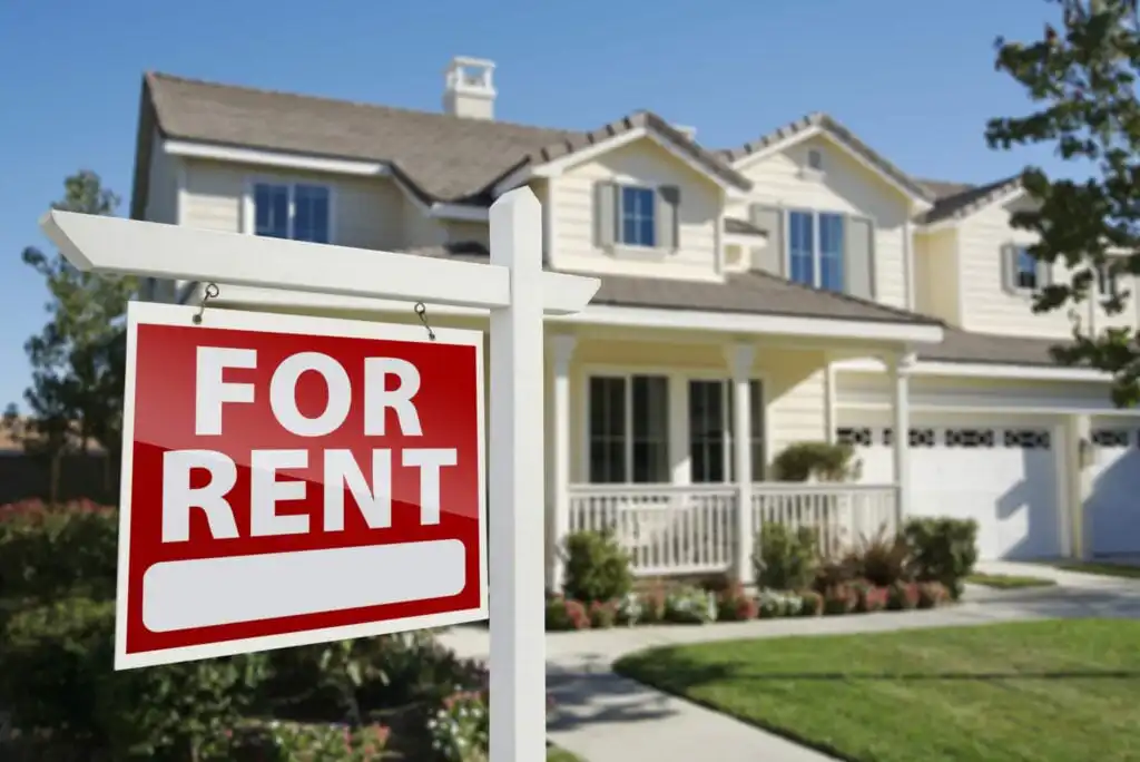 A large For Rent sign is displayed in front of a modern two-story beige house with white trim, a porch, manicured lawn, and landscaped yard on a bright, sunny day.