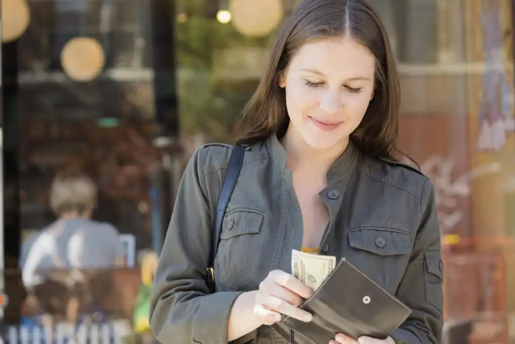 A woman with long brown hair smiles while taking cash out of her black wallet outdoors, with a blurred background of a storefront and people.