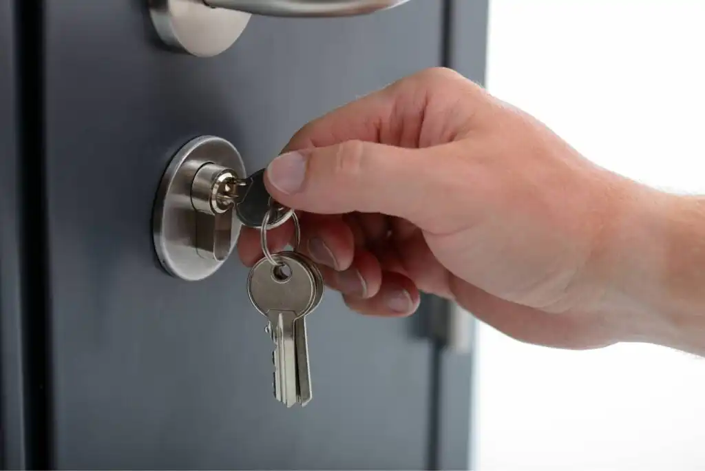 A hand holding a key ring is inserting a key into the lock of a dark-colored door, with a metallic door handle visible above the lock.