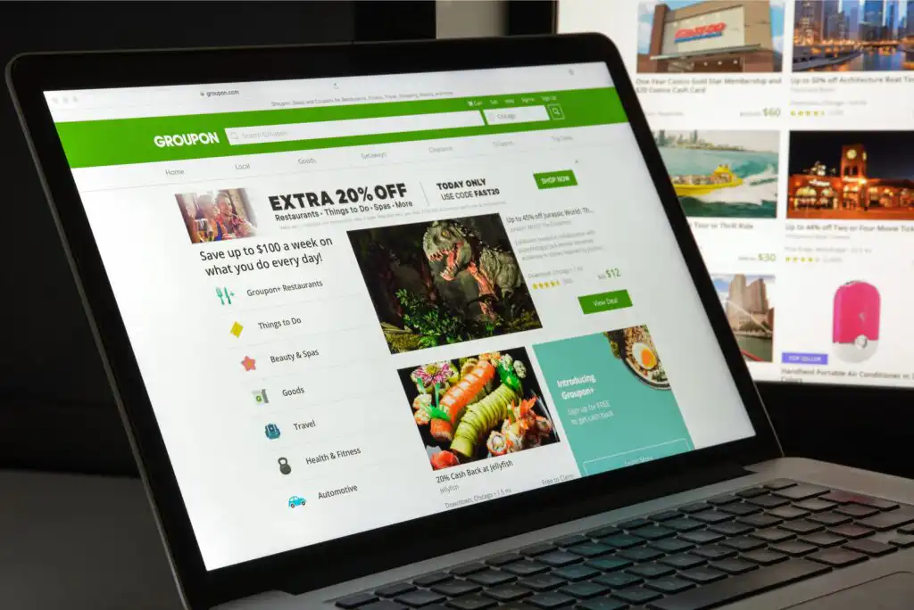 A laptop screen displays the Groupon website, showing deals and discounts on various categories such as restaurants, goods, travel, and health. The site features images of food, animals, and discount offers.